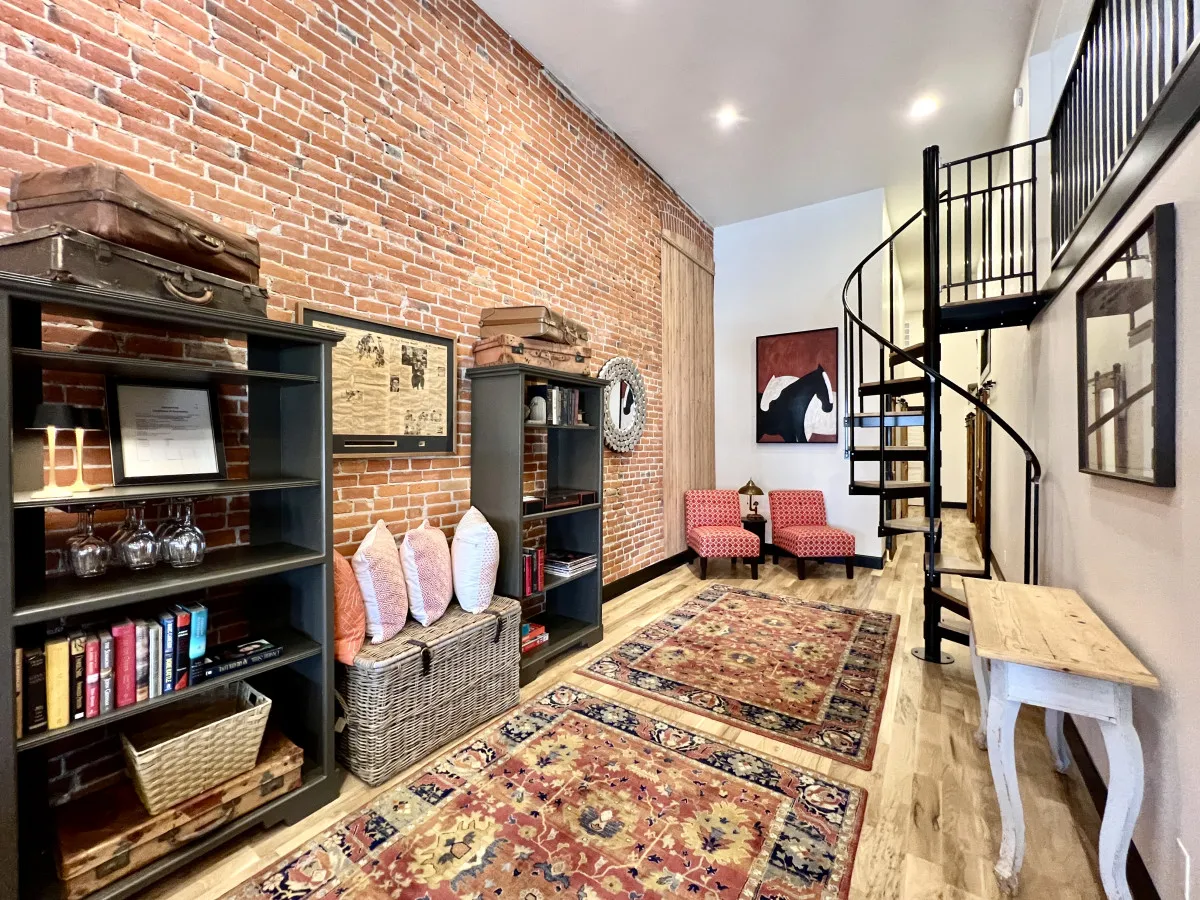 A vibrant, eclectic living area with exposed brick walls, a spiral staircase, bookshelves filled with books and decor, comfortable accent chairs, and a colorful rug. The space is bright, airy, and designed for both socializing and quiet work or reading breaks.