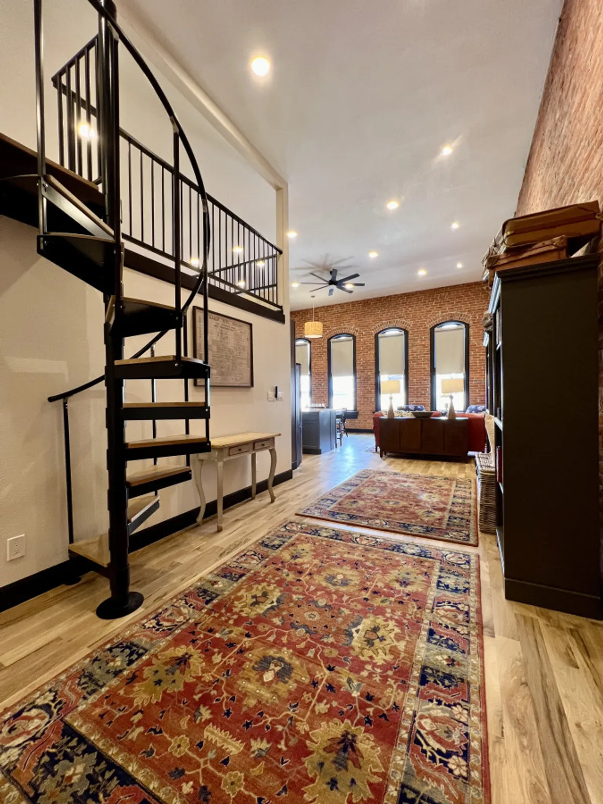 The photo shows a spiral staircase leading to a lofted area with brick walls, large windows that flood the space with natural light, and traditional rugs. The space feels open and inviting, offering glimpses of both work and leisure areas.