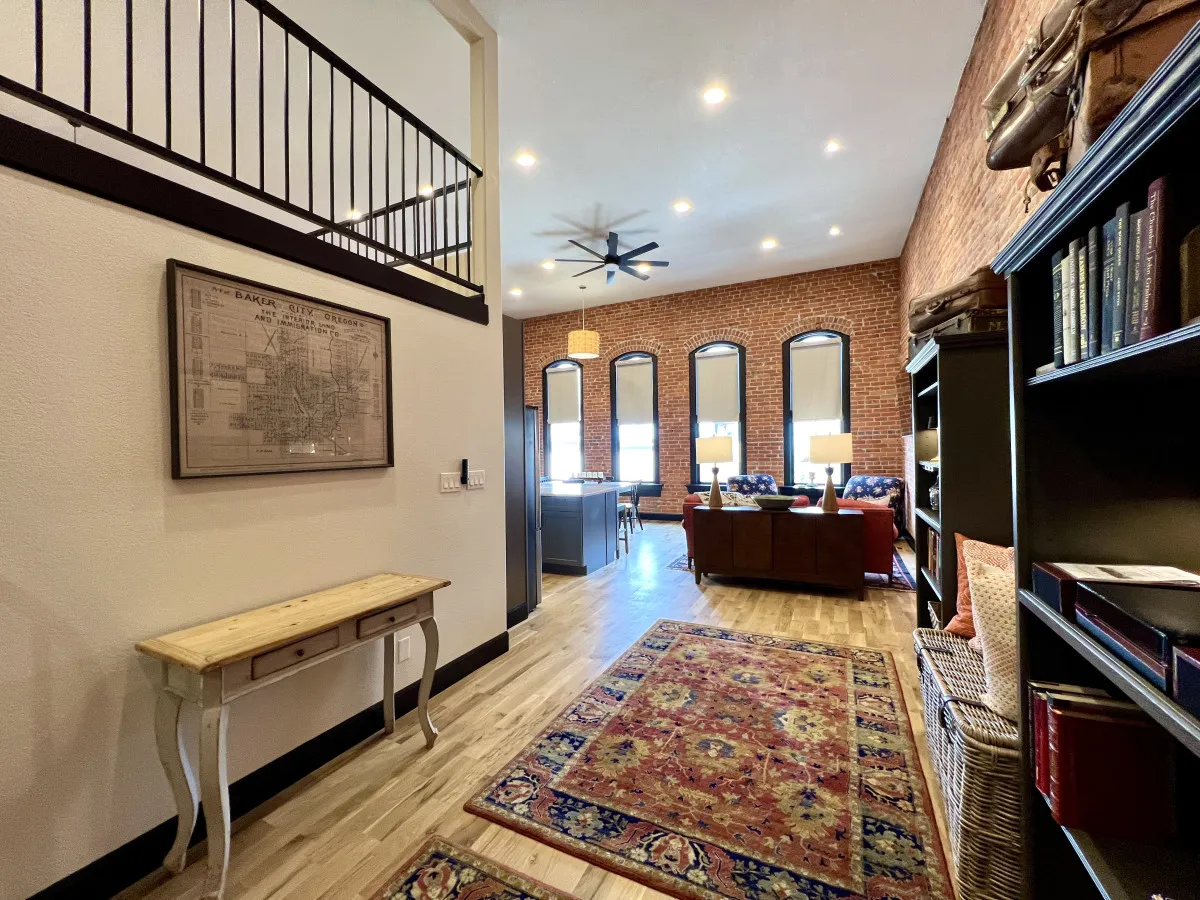 A spacious living area bathed in natural light from large windows, featuring an exposed brick wall, plush and comfortable seating, a decorative rug, and a bookshelf. The space seamlessly combines relaxation and socializing, with views out to Main Street below.