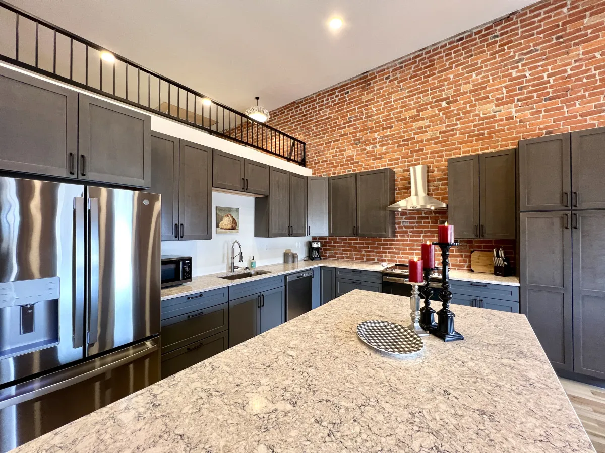 A sophisticated kitchen area with stainless steel appliances, granite countertops, and dark cabinetry set against an exposed brick wall. The space is designed for entertaining, with plenty of room for preparing meals or enjoying breakfast together.