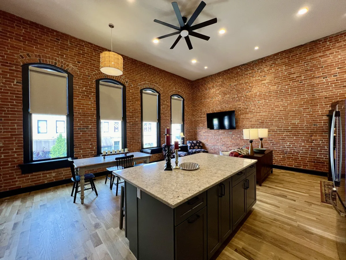 A cohesive open-plan environment featuring a plush sofa area, an island counter, brick walls, statement lighting, and large windows that flood the space with natural light and offer vibrant views of downtown Baker City.