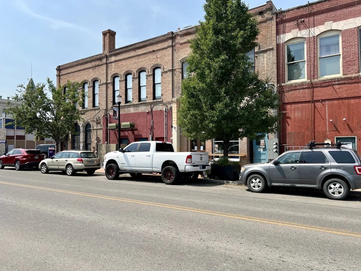 A vibrant exterior shot of a historic brick building located downtown, framed by a leafy green tree and several parked cars. The view captures the lively street atmosphere and hints at the various shops, dining spots, and walkable amenities nearby.