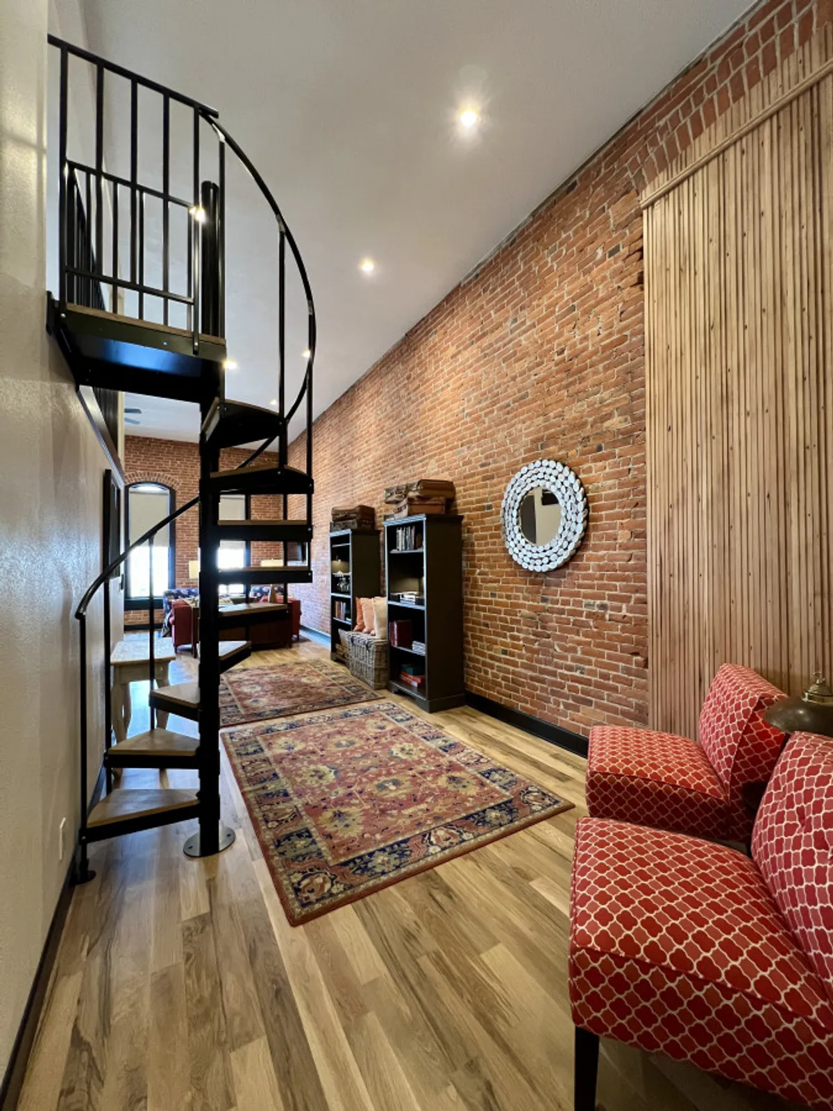 This image features a spiral staircase leading up to a lofted area, with red patterned chairs, bookshelves, a decorative mirror, and an exposed brick wall. There's ample natural light, suggesting an open view from the loft over the main living space, and the arrangement hints at both a productive and relaxing environment.
