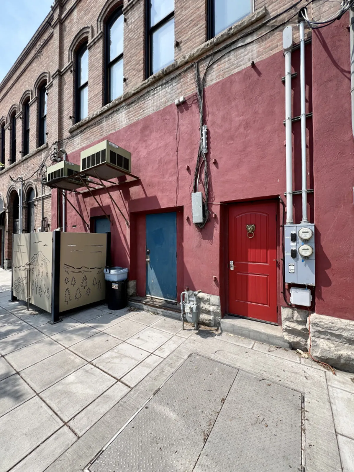 This exterior image depicts the building’s façade featuring a distinctive red and blue door, air conditioning units, and a decorative panel with nature illustrations. The scene is set in an urban environment, with street-level details that hint at the building's central location.