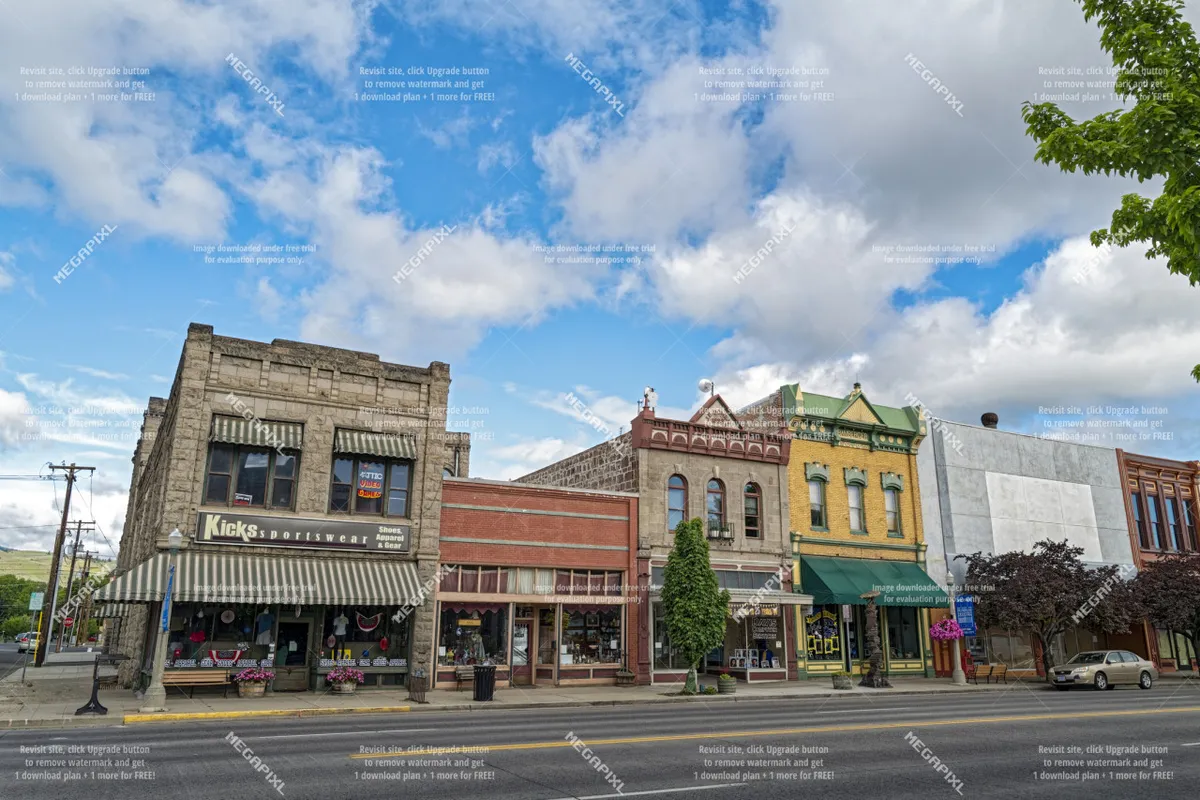 A cheerful streetscape in downtown Baker City, highlighting colorful building facades, street trees, and local storefronts including a sportswear shop. People are seen strolling or visiting shops, suggesting a vibrant local scene.
