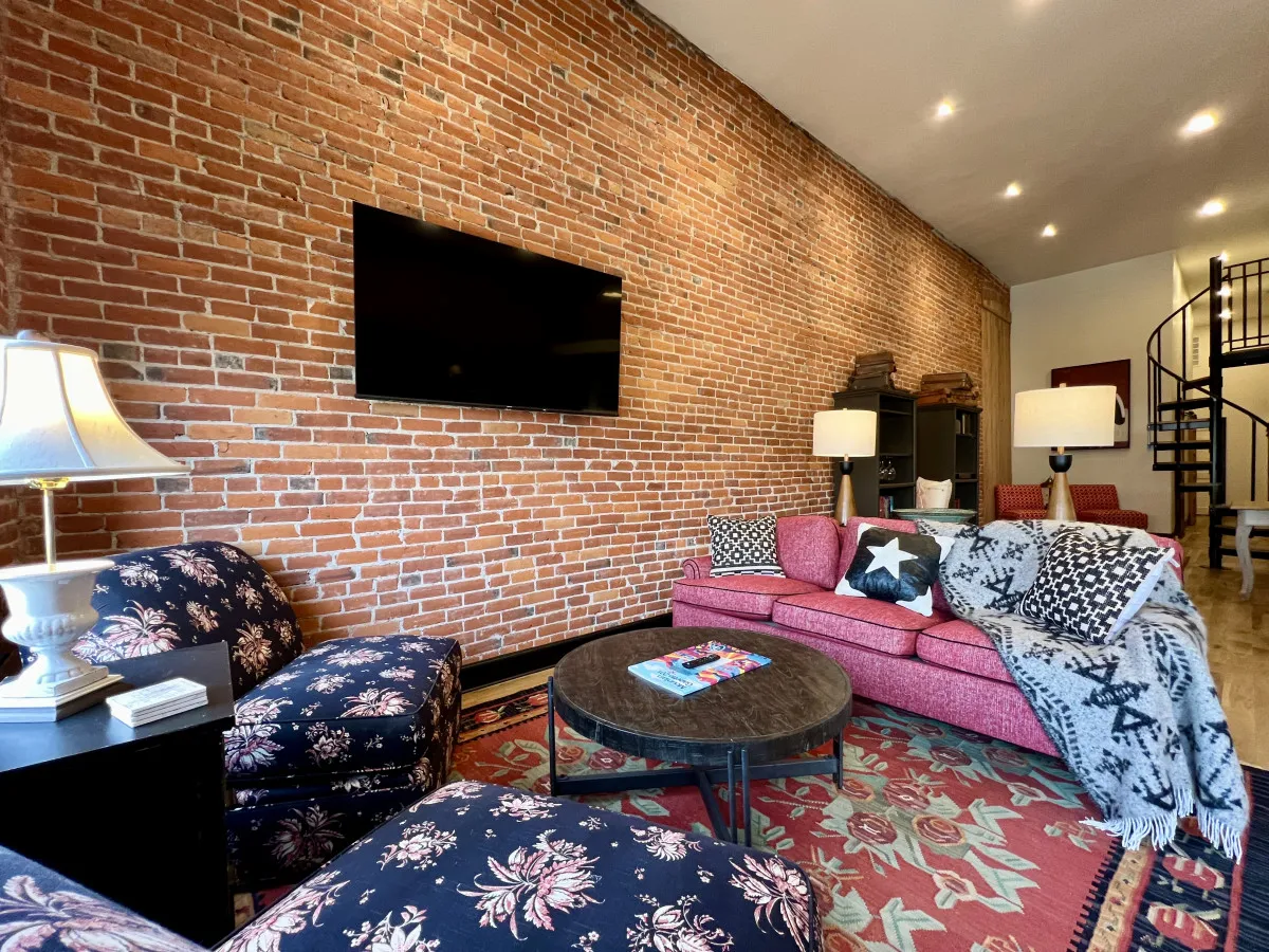 A welcoming living area featuring a stylish brick wall, plush sofa with decorative pillows, a modern coffee table, and soft lighting from a contemporary lamp. The ambiance suggests a cozy setting perfect for relaxing, reading, or socializing with friends and family.