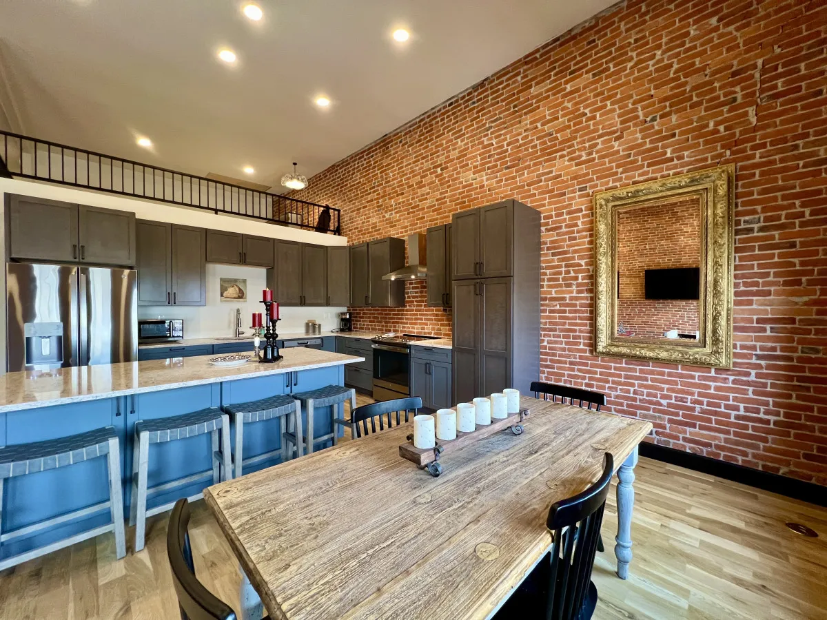 The image showcases a stylish, light-filled downtown loft interior featuring a striking exposed brick wall, a sleek modern kitchen with stainless steel appliances, a dining table set with elegant candles, and a large ornate mirror reflecting the inviting space. The room blends contemporary comforts with historic character, creating a welcoming setting for both intimate dinners and lively gatherings.