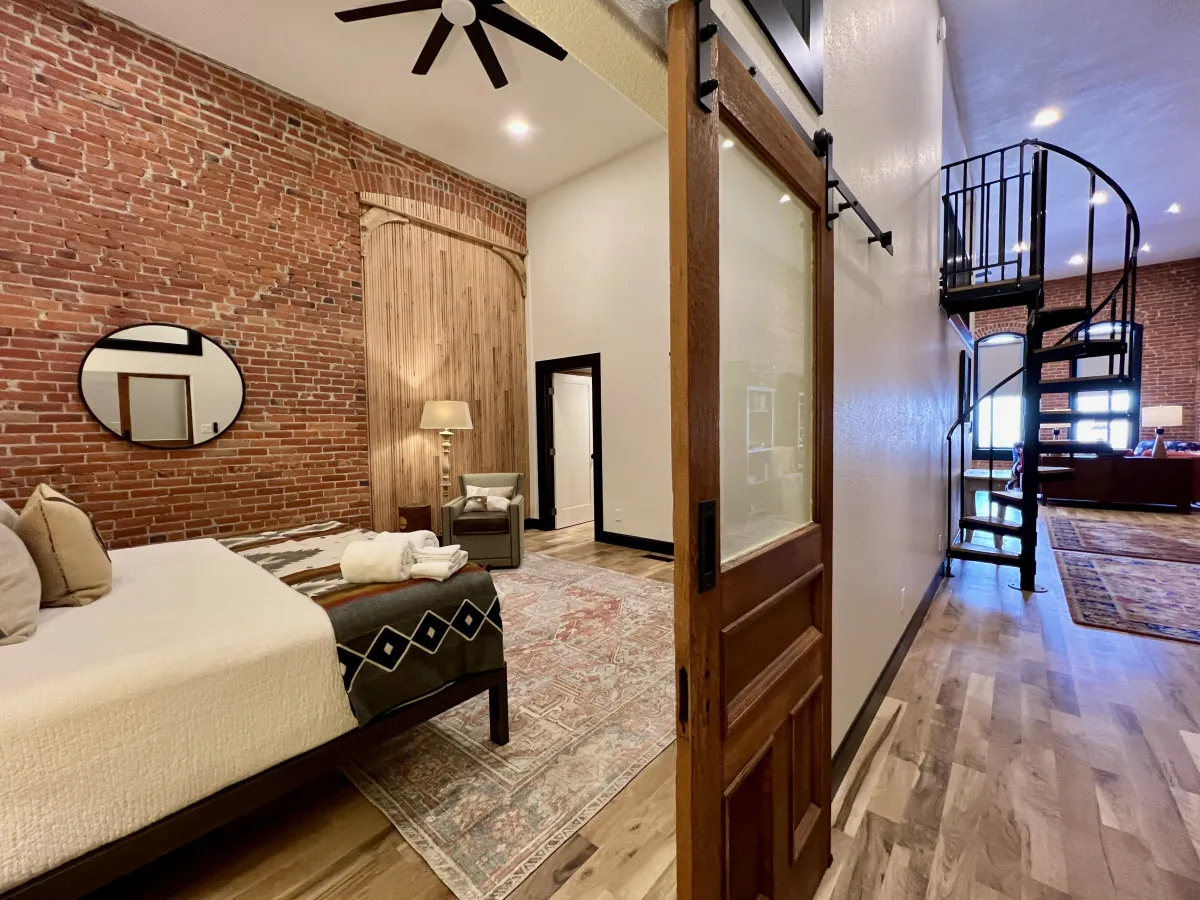 A cozy nook featuring a spiral staircase, a large round mirror, exposed brick wall, and a stylish Murphy bed setup with decorative pillows. There's a sense of comfort and flexibility, ideal for work, sleep, or quiet moments.