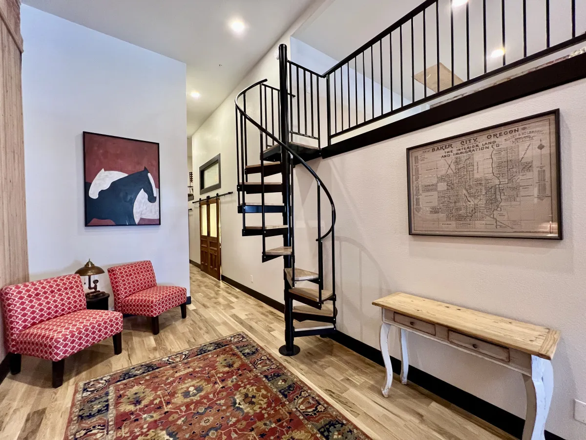 An interior entryway featuring a spiral staircase, red patterned chairs, artwork, a wooden console table, and a vintage map. The area feels both welcoming and stylish, suggesting a space that blends comfort with historical charm and creative inspiration.